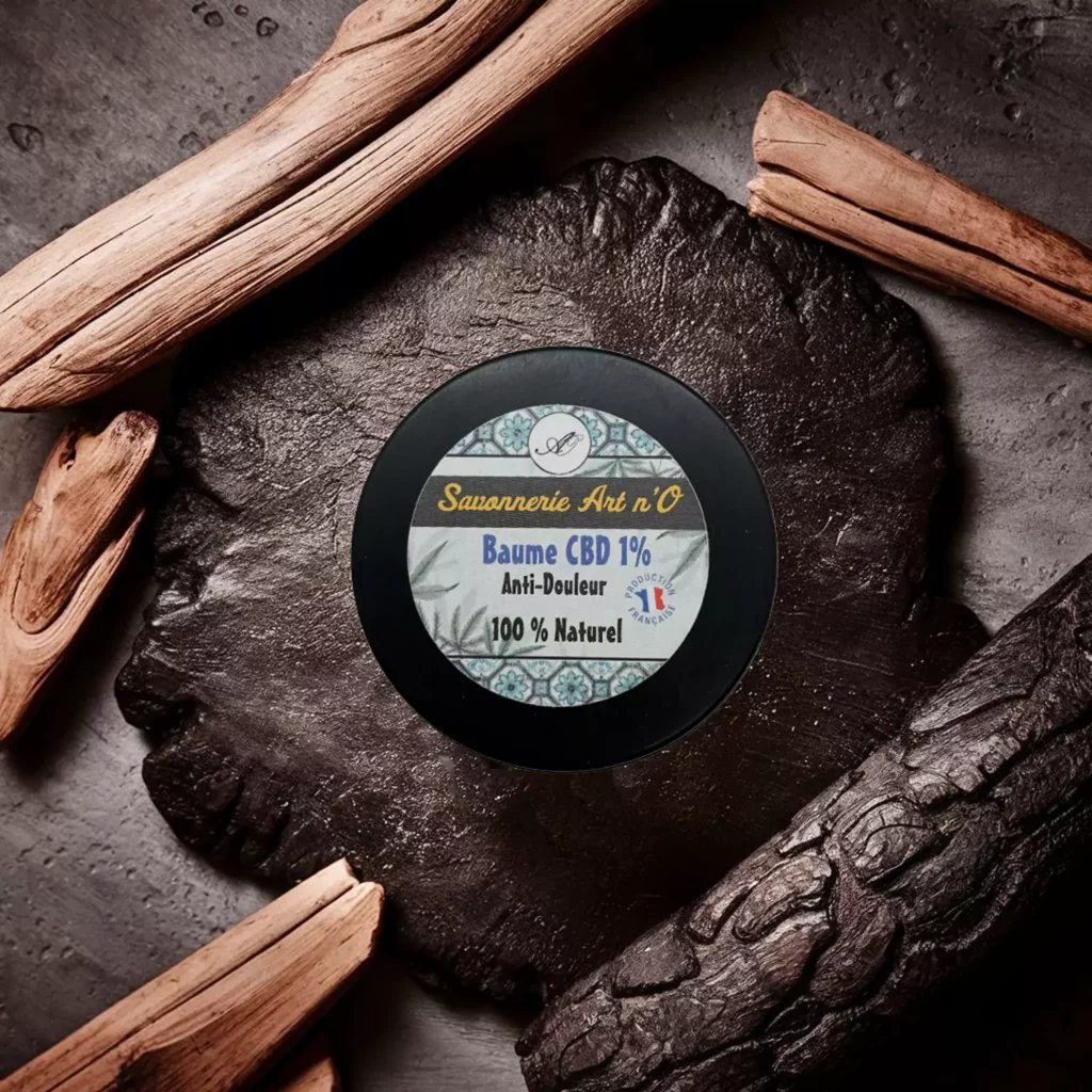Baume 1% soulage douleur de Savonnerie Art n’O, petit pot noir posé sur pierre, entouré de morceaux de bois.