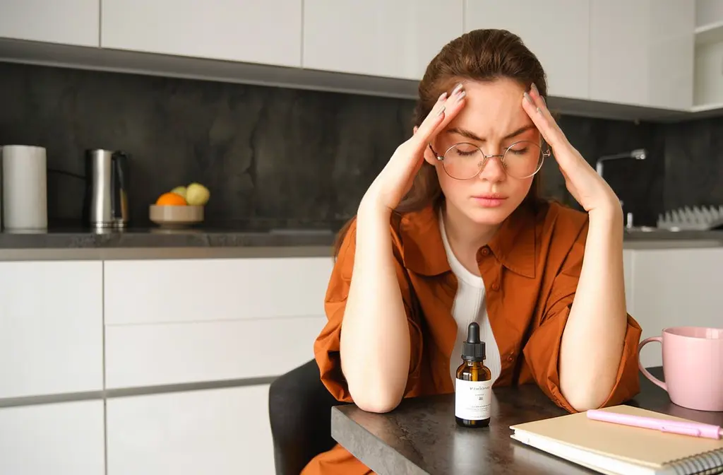 Femme au visage douloureux tenant ses tempes, bouteille d’huile essentielle sur la table dans une cuisine moderne.