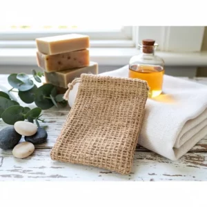 Pochette en sisal exfoliante posée sur serviette blanche, avec savon en bloc, galets et huile essentielle en arrière-plan.
