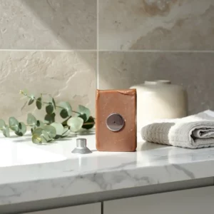 Porte-savon magnétique en savon brun posé sur plan de salle de bain, with ventouse métallique et accessoire, près d’une serviette et d’un vase.